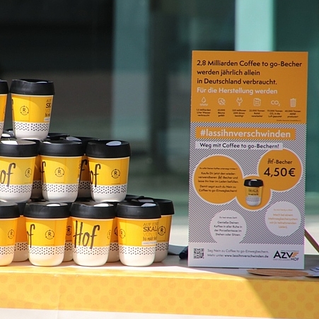 Mehrere Mehrweg- Coffee to go-Becher (HofBecher) stehen auf einem Tisch. Daneben ist ein Schild mit Informationen zur Kampagne #lassihnverschwinden