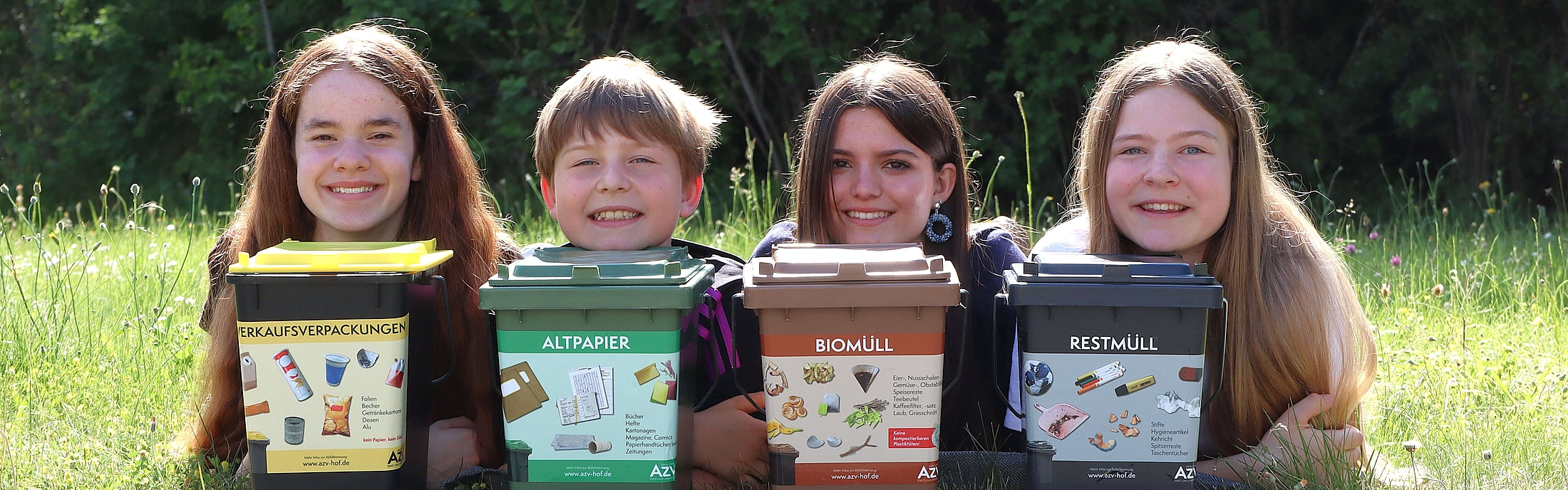Drei Kinder auf einer Wiese mit jeweils einer kleinen Mülltonne. Bioabfall, Restmüllabfall und Papierabfall.