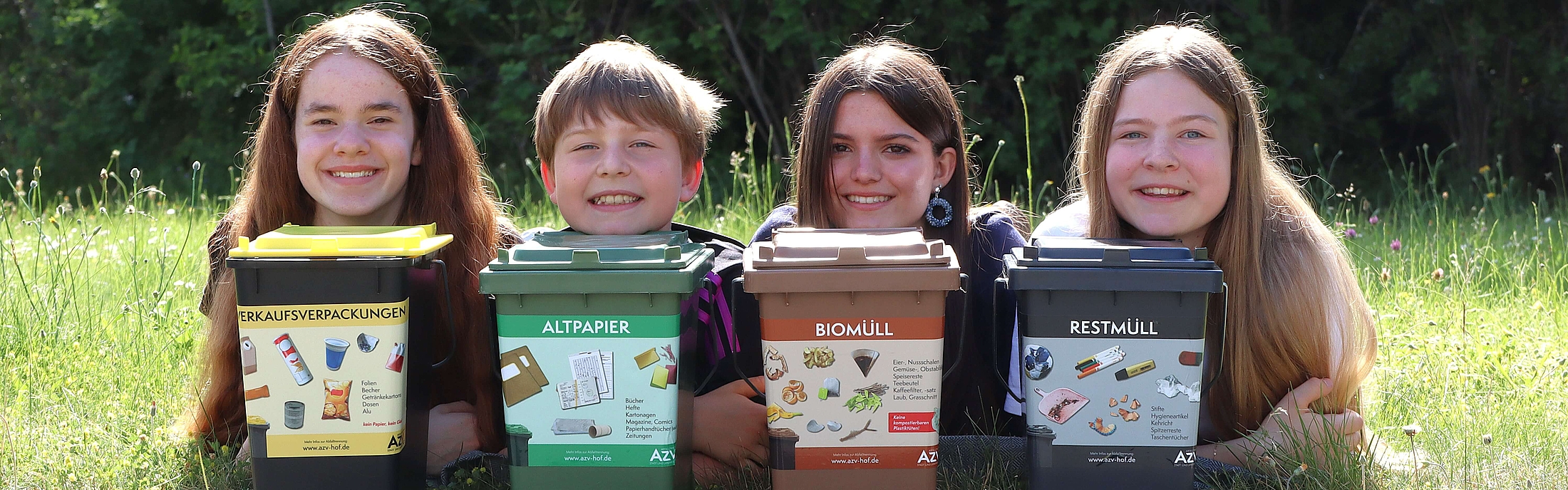 Drei Kinder auf einer Wiese mit jeweils einer kleinen Mülltonne. Bioabfall, Restmüllabfall und Papierabfall.
