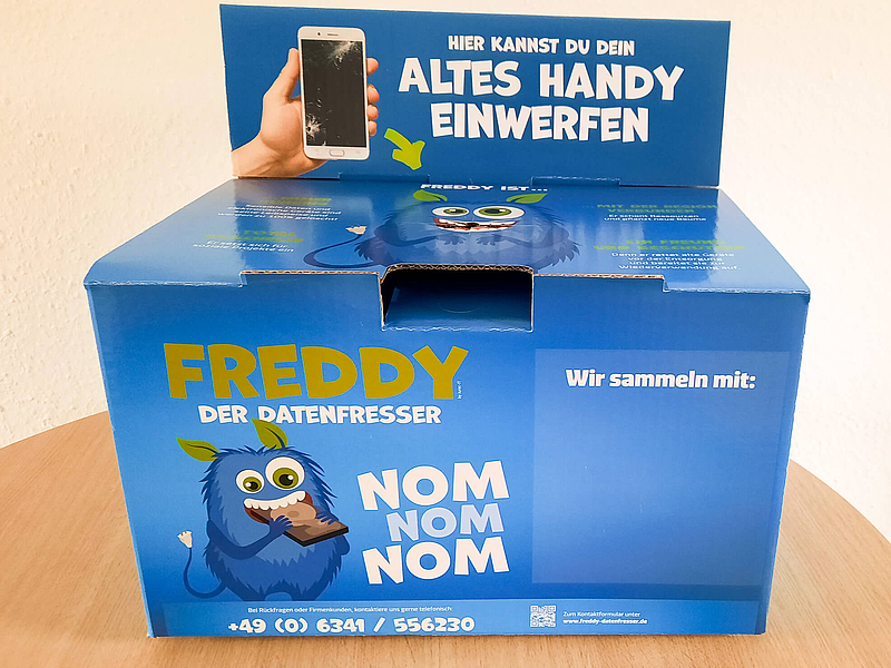 Handykiste zum Sammeln von unbenutzten und unbrauchbaren Handys. Der blaue Faltkarton liegt auf einem Holztisch. Bedruckt ist der Karton mit einem blauen Monster "Freddy Datenfresser".